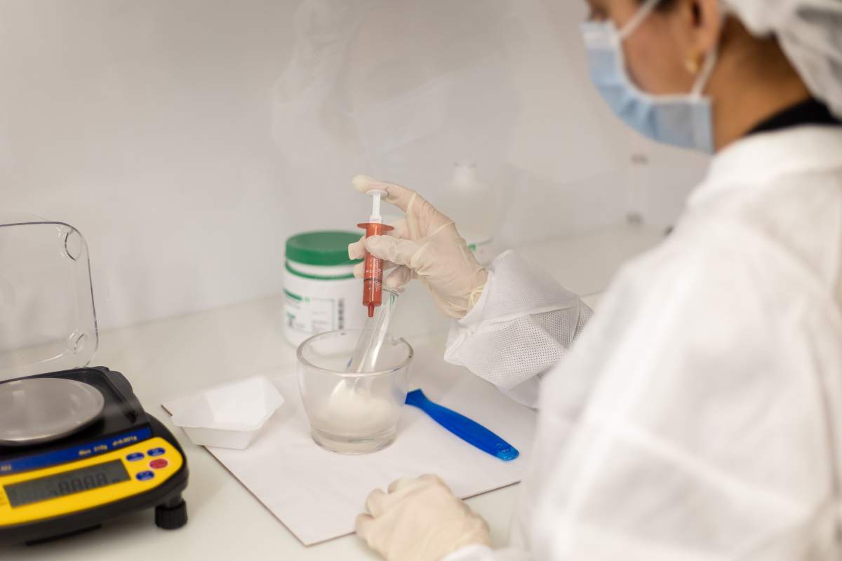 Pharmacist wearing protective gear, including a mask, gloves, and lab coat, carefully mixing ingredients in a glass bowl for compounding medication.